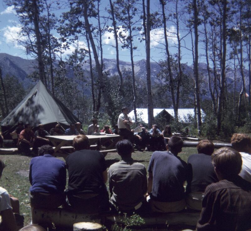A group of people are gathered outdoors, seemingly at a campsite or retreat. They are sitting on logs and benches, facing a speaker who is standing near a picnic table. A large tent is visible on the left side of the frame, and trees surround the gathering, with mountains in the background. The atmosphere appears relaxed and informal.